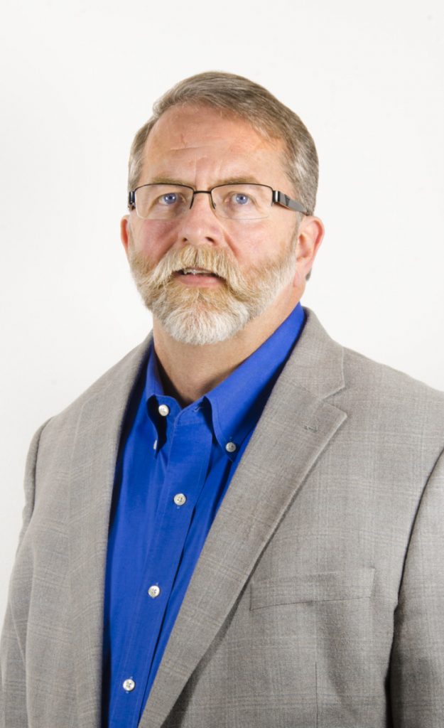 A middle-aged man with short, light brown and gray hair, a full mustache and beard, wearing glasses, a gray suit jacket, and a blue shirt, posed against a plain white background.