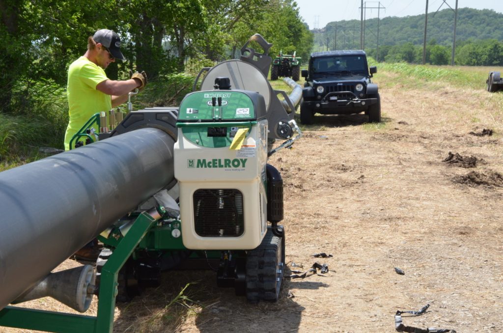 A worker in a neon yellow shirt operates a McElroy machine to weld large black pipes outdoors, with trees on the left and a parked black Jeep on the dirt road nearby.