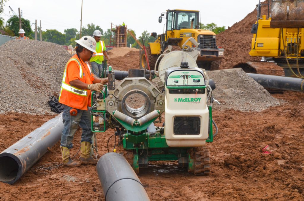 A construction worker wearing a safety vest and helmet operates a large pipe fusion machine on a muddy construction site, with heavy machinery and pipes visible in the background.
