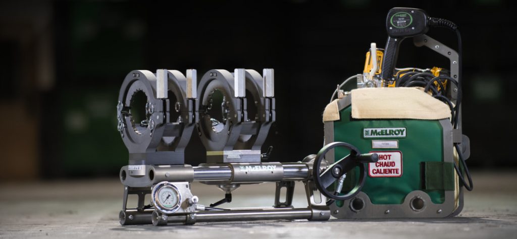 A McElroy pipe fusion machine with three metal clamps, a pressure gauge, and a green control unit labeled HOT CHAUD CALIENTE, set on a concrete floor with a dark, blurred background.