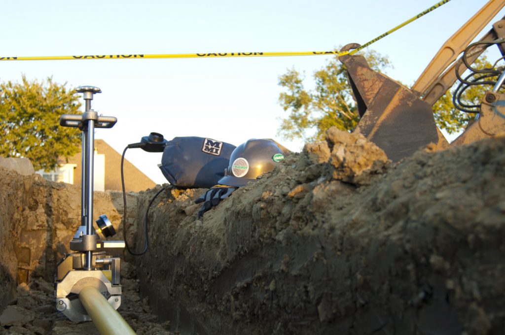 A close-up of excavation work with caution tape, machinery, and equipment on a construction site. A metal tool is attached to a yellow pipe in a trench with soil, and a helmet rests nearby. Trees and houses are visible in the background.