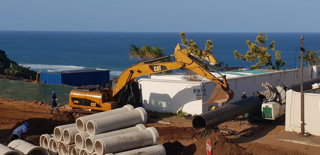 An excavator lifts a large black pipe at a coastal construction site, with workers nearby and concrete pipes on the ground. The ocean and blue sky are visible in the background.