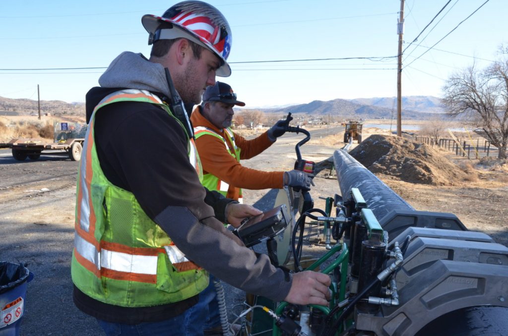 Two construction workers in safety vests and helmets operate machinery to work on a pipeline by a rural roadside; dry landscape and mountains are visible in the background.