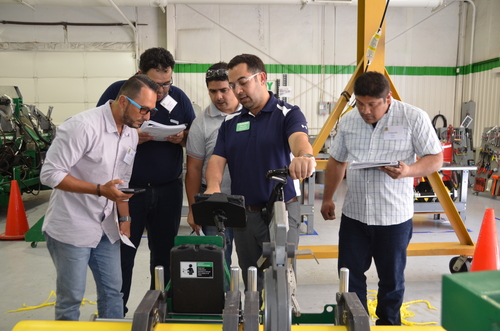 Five men in casual shirts stand around a piece of industrial equipment in a workshop, examining it closely and taking notes. Orange cones and machinery are visible in the background.