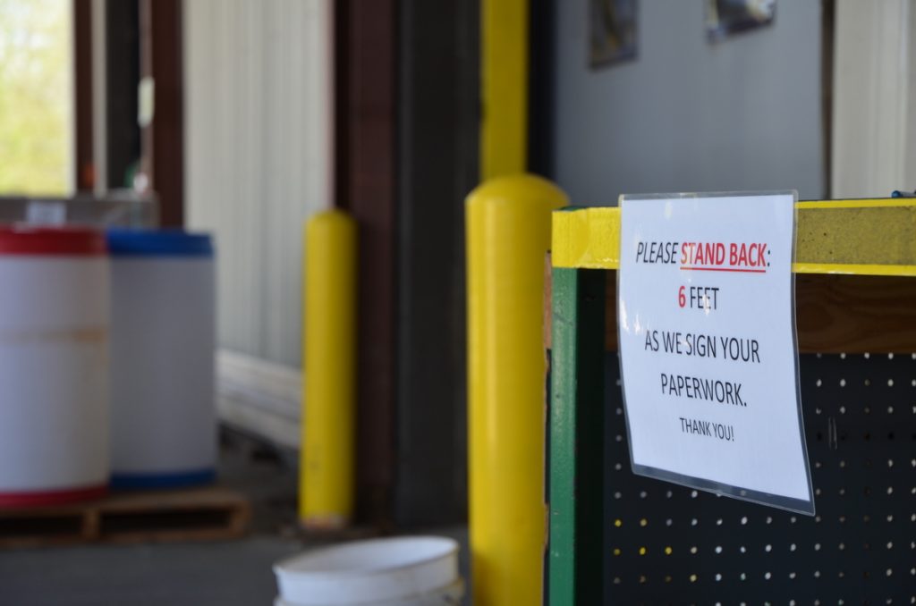 A sign on a counter reads, Please stand back: 6 feet as we sign your paperwork. Thank you! Large barrels and yellow safety bollards are visible in the background, suggesting an industrial or warehouse setting.