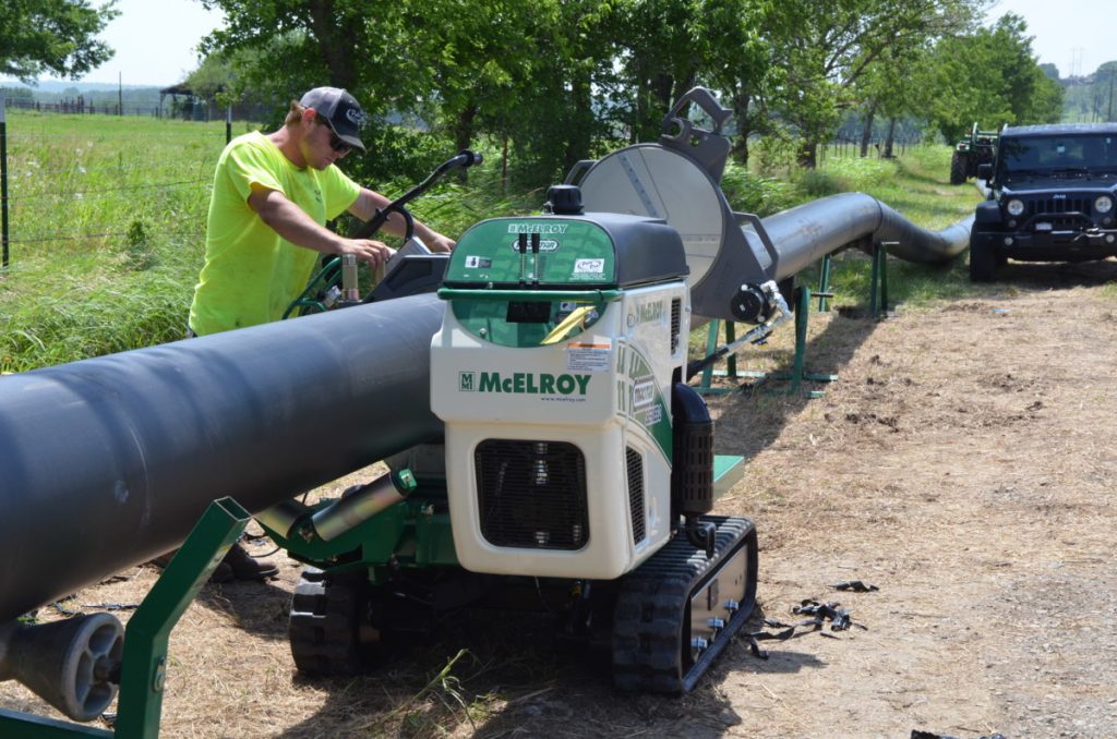 A worker in a yellow shirt operates a McElroy machine to fuse large black pipes together outdoors, with trees, grass, and a black vehicle visible in the background on a sunny day.