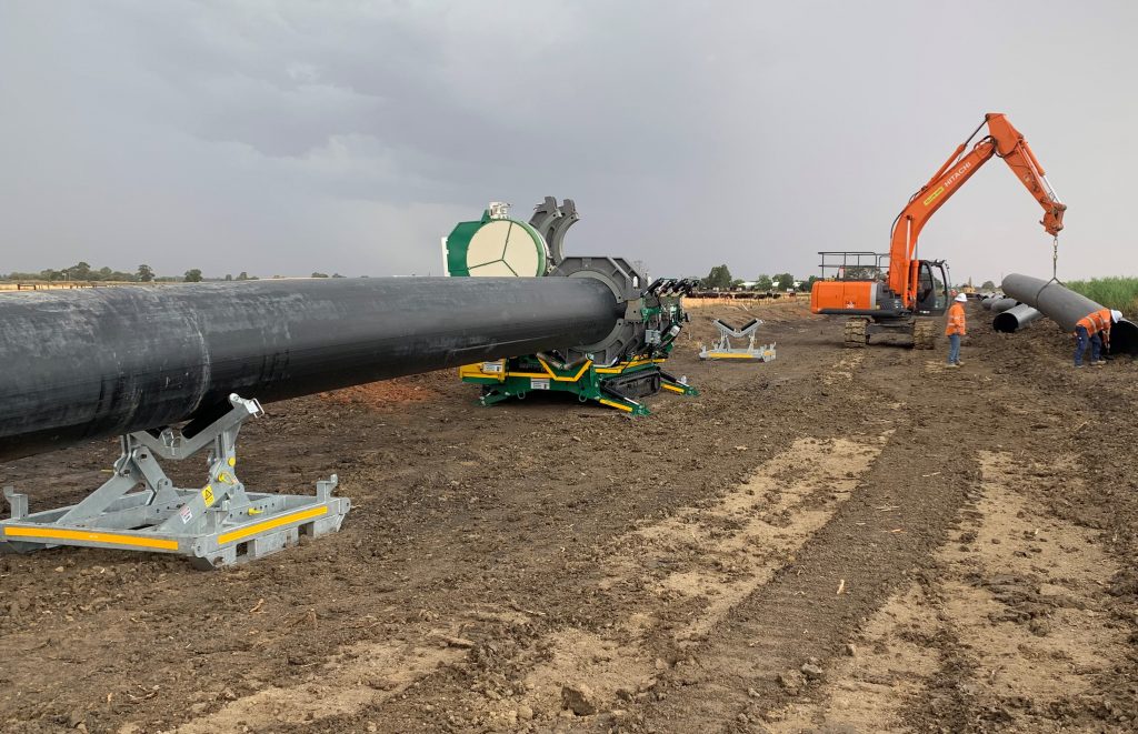 An excavator and workers are installing large black pipes on a dirt construction site under a cloudy sky. Equipment and machinery are positioned along the pipeline.