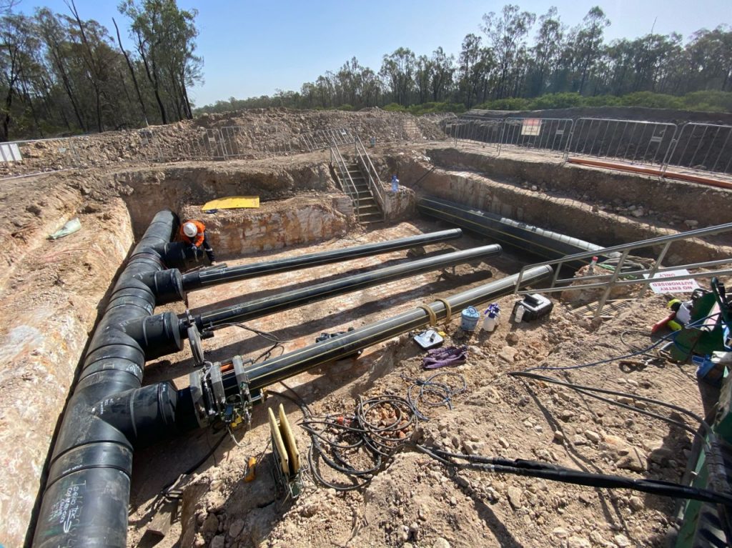 A construction site showing large black pipes being installed underground, with workers in safety gear nearby and trees in the background. Tools and equipment are scattered around the excavation area.