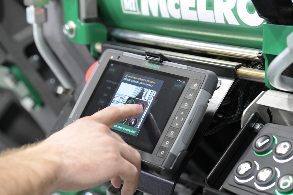 A person’s hand operates a touchscreen control panel attached to industrial machinery, displaying technical diagrams and interface elements. The equipment and controls are branded with the McElroy logo.