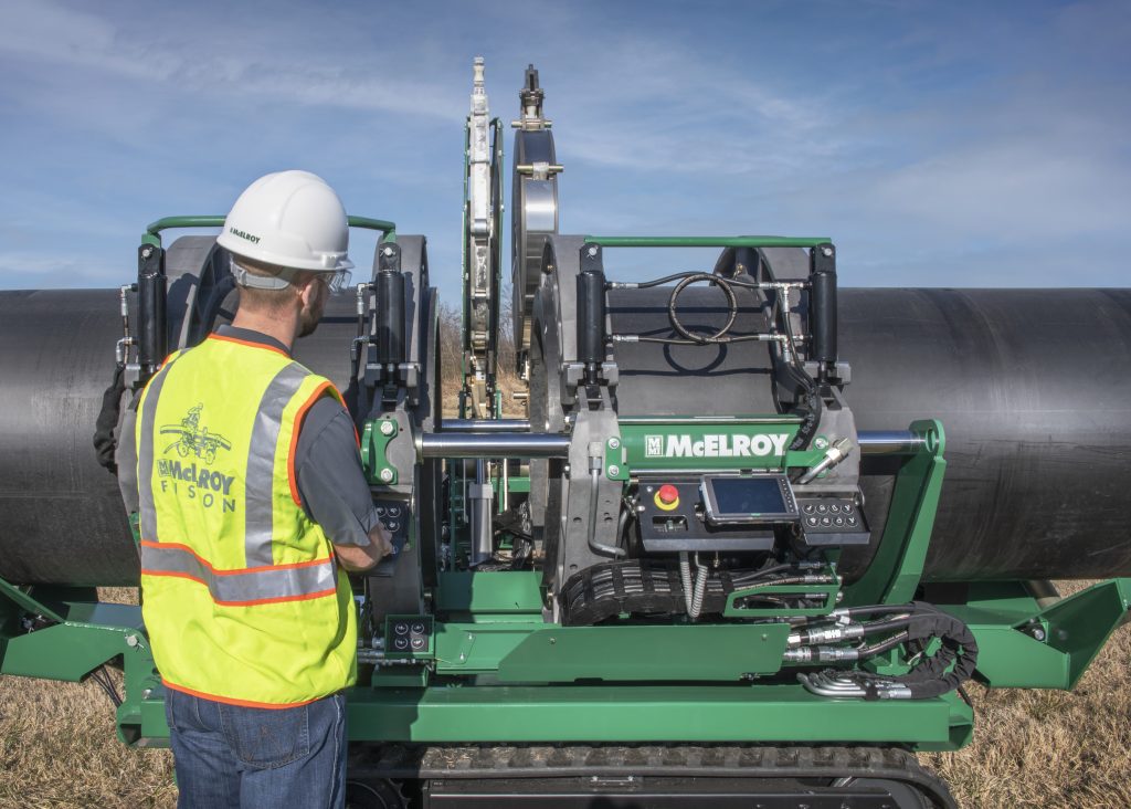 A worker in a hard hat and safety vest operates a McElroy fusion machine on large black pipes outdoors under a blue sky.