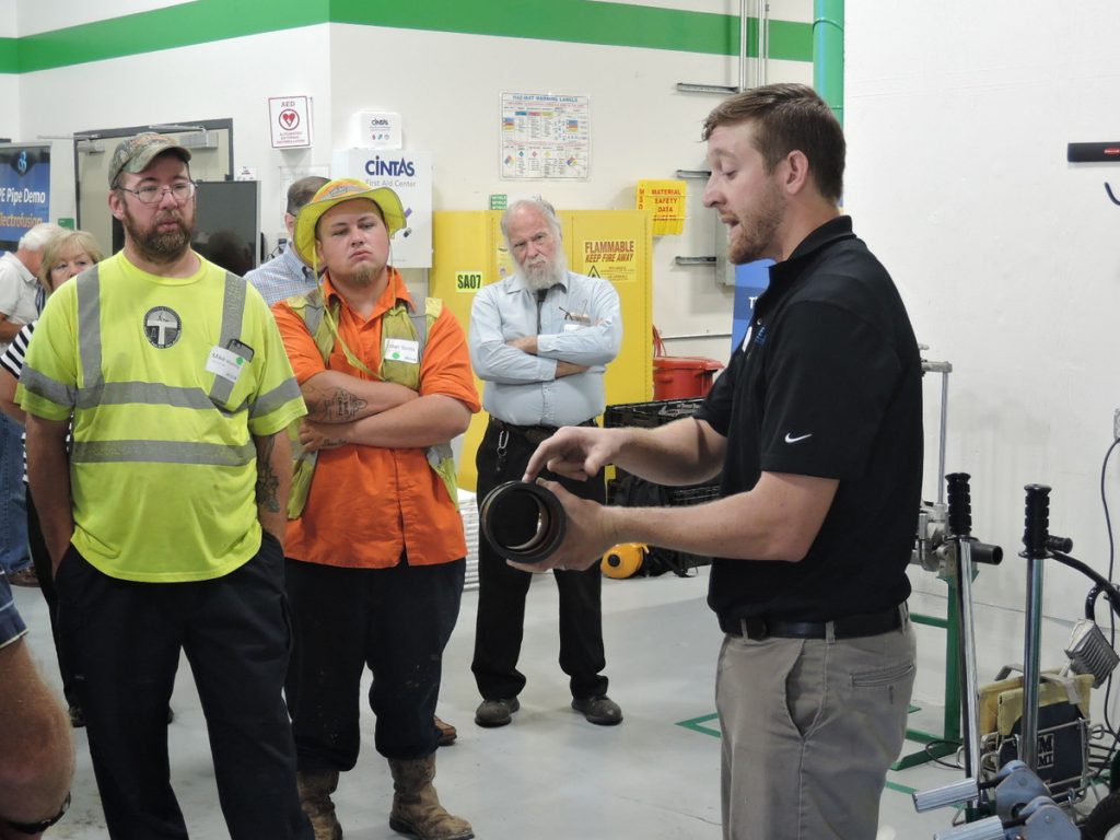 A man in a black shirt demonstrates a pipe fitting to a group of workers in safety gear inside an industrial facility, while others watch attentively. Equipment and safety signs are visible in the background.