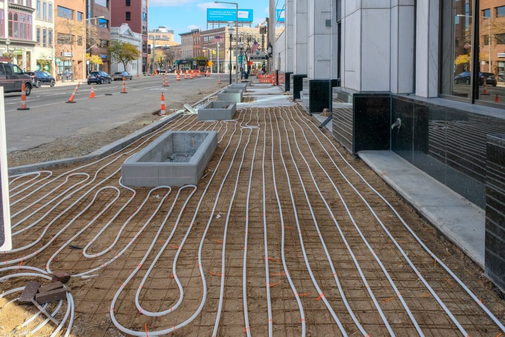 White pipes arranged in loops are laid out on the ground along a city sidewalk under construction, with concrete planters and orange safety cones visible by the street and buildings in the background.