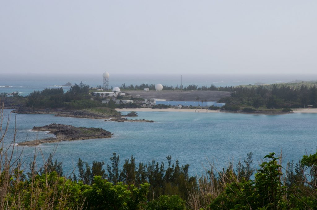 A calm bay with turquoise water, sandy beaches, small rocky islands, and a facility with several white radar domes and antennas surrounded by trees in the background.