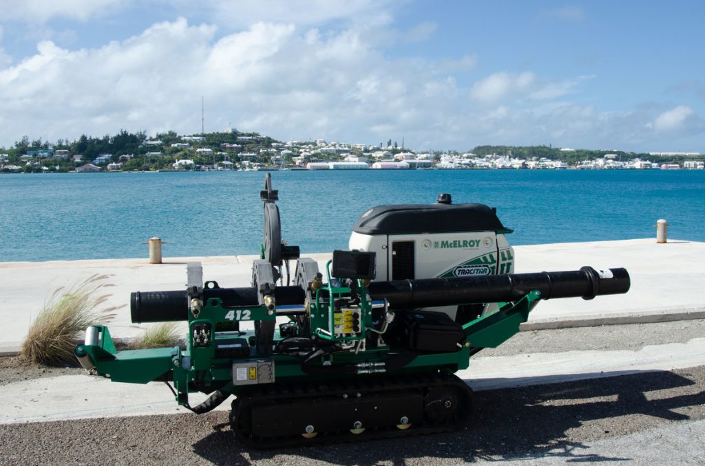 A green pipe fusion machine labeled McElroy sits on a concrete dock by blue water, with a coastal town and greenery visible in the background under a partly cloudy sky.