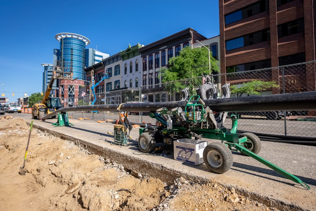 A large machine works on installing a black pipeline along a city street, with construction equipment, buildings, and fencing visible under a clear blue sky.