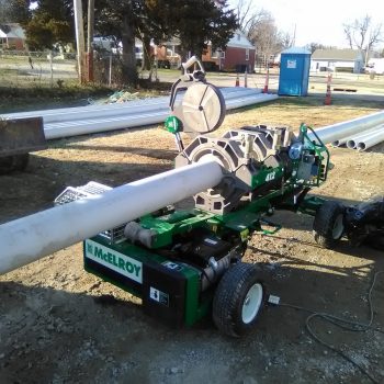 A green McElroy fusion machine aligns and fuses long sections of white plastic pipe at an outdoor construction site, with pipes, equipment, and a portable toilet visible in the background.