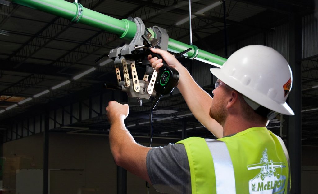 A construction worker in a hard hat and safety vest operates a fusion machine to join green plastic pipes inside an industrial building.
