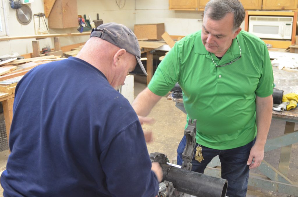 Two men work together in a workshop; one wears a green shirt and watches as the other, in a blue shirt, operates a tool on a black pipe. Woodworking equipment and cabinets are visible in the background.