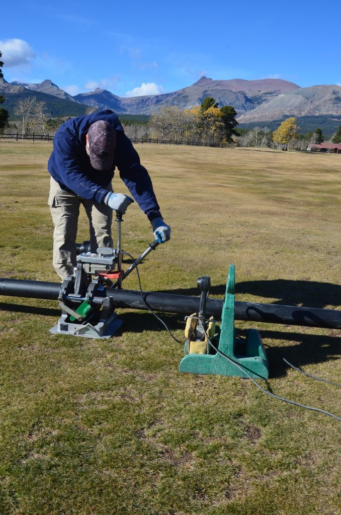 A person in outdoor clothing operates a machine on a grassy field, working with a long black pipe. Trees and mountains are visible in the background under a blue sky.