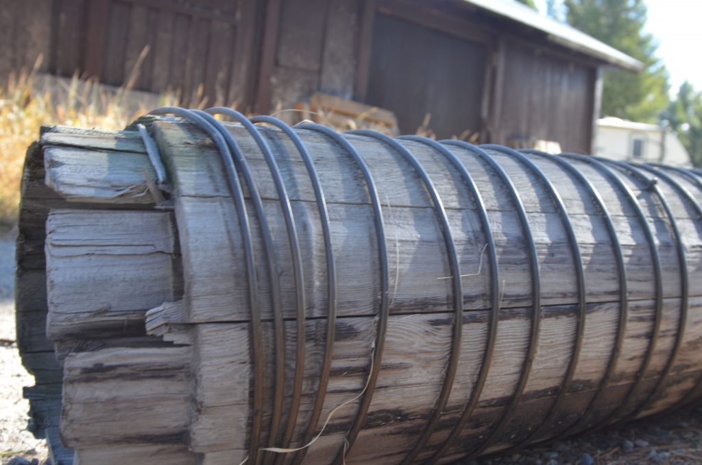 A close-up shot of a large weathered wooden log wrapped in metal wire, lying horizontally outdoors near a rustic building with blurred background.