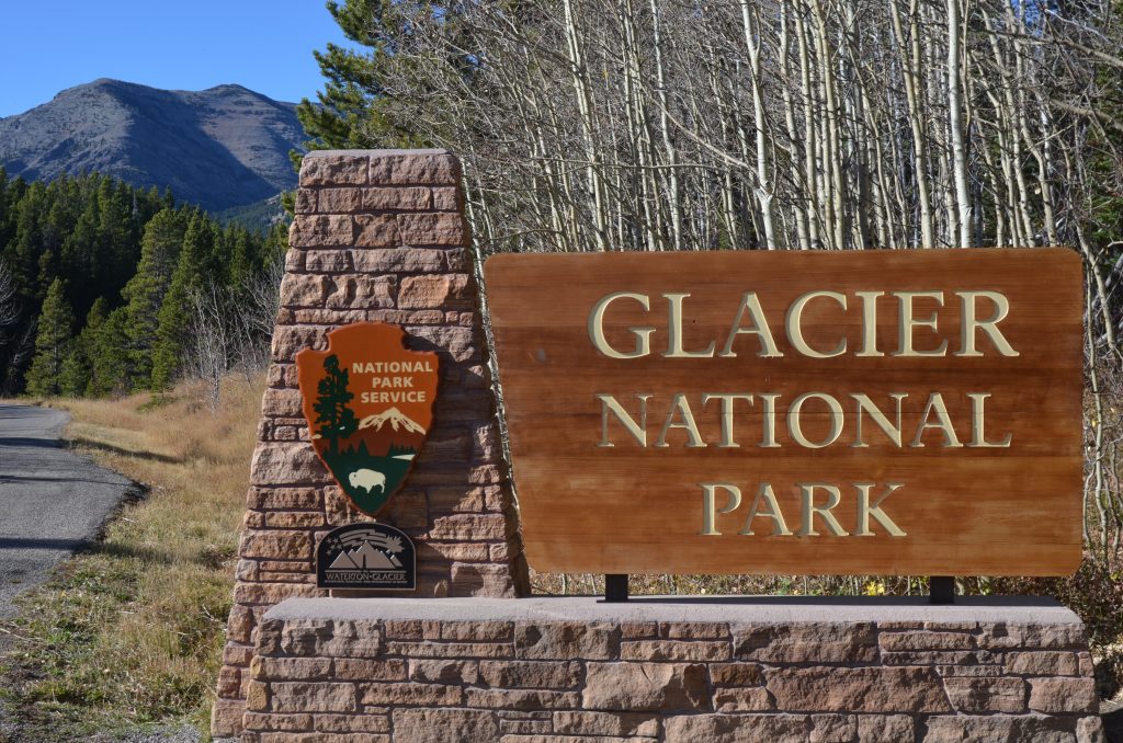 A large wooden sign reads Glacier National Park next to a stone pillar with a National Park Service emblem, with trees and mountains in the background under a clear blue sky.