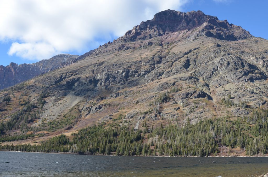 A rocky, rugged mountain rises behind a tree-lined shore and a rippling lake under a partly cloudy sky.