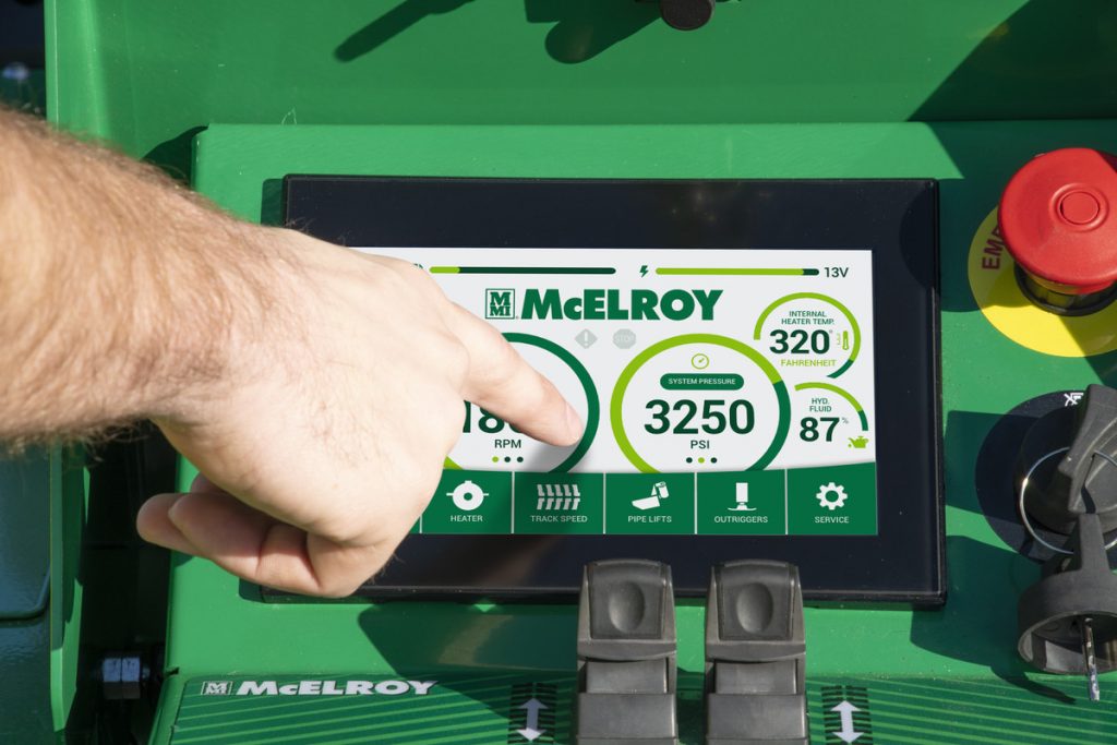 A person’s hand points at a touchscreen display on a green McElroy machine, showing data such as RPM, PSI, hydraulic fluid temperature, and other operational metrics.