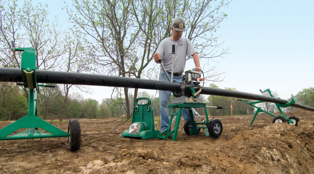 A worker in a hard hat operates machinery to fuse sections of a large black pipe outdoors on a dirt surface, with trees and green equipment visible in the background.