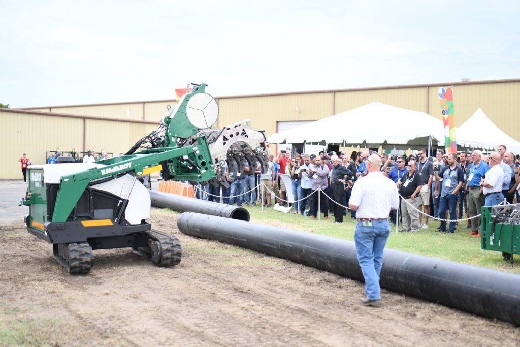 A crowd watches as a green industrial machine prepares to lift a large black pipe outdoors near a building and white tents during a demonstration event.