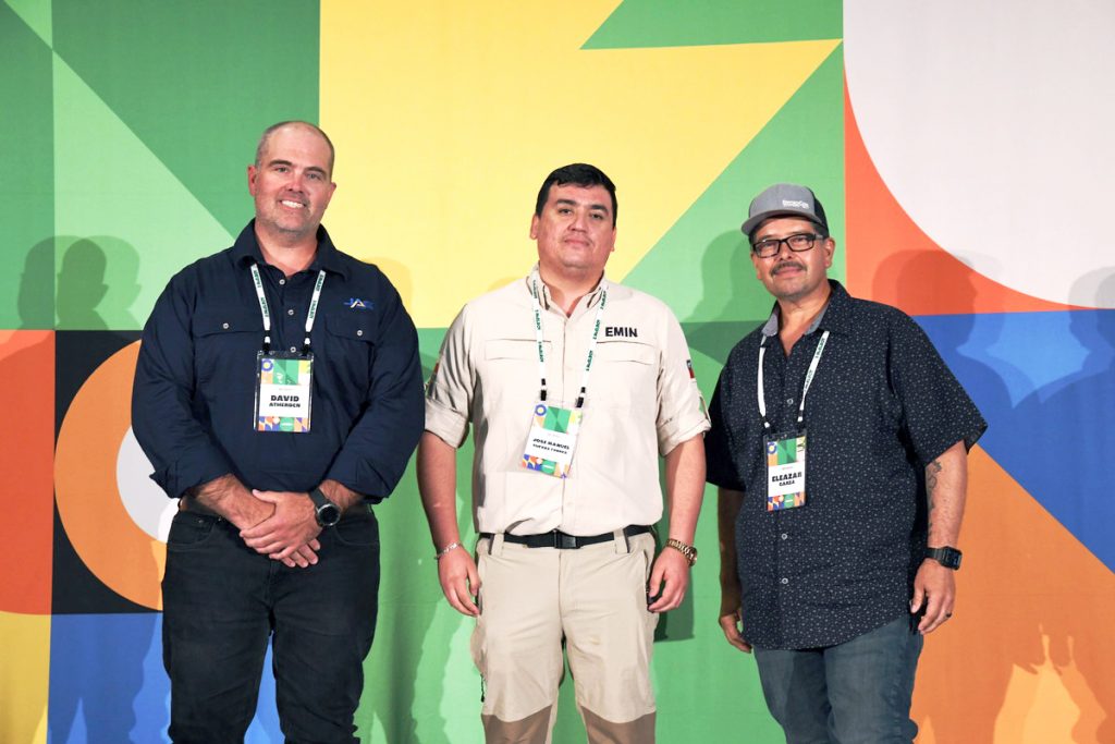 Three men stand side by side, smiling at the camera, wearing conference badges against a colorful geometric background. The man on the left wears a dark shirt, the middle man wears beige, and the man on the right wears a cap and glasses.