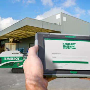 A person holding a tablet displaying the McElroy Equipment Inspection login screen in front of an industrial building with McElroy equipment outside under a sunny sky.