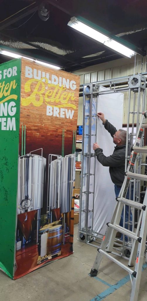 A man assembles a metal display frame next to a colorful booth panel with the text Building Better Brew and images of large brewing tanks in an industrial indoor setting.