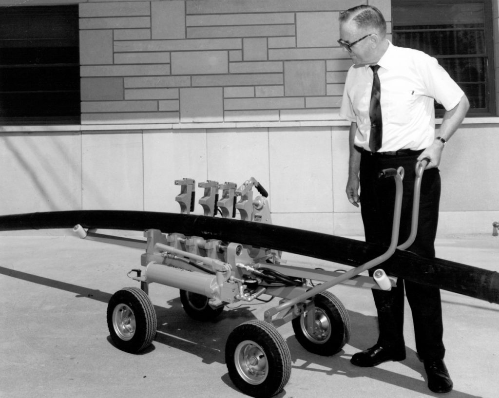 A man in a white shirt and tie stands next to a device on four wheels, holding its handle. The device appears to be designed for manipulating or transporting a long, curved object.
