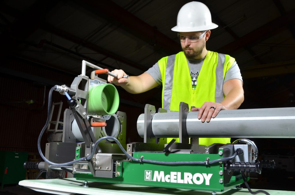 A worker wearing a white hard hat, safety glasses, and a yellow safety vest operates a McElroy pipe fusion machine, aligning a green pipe with a large gray pipe indoors.