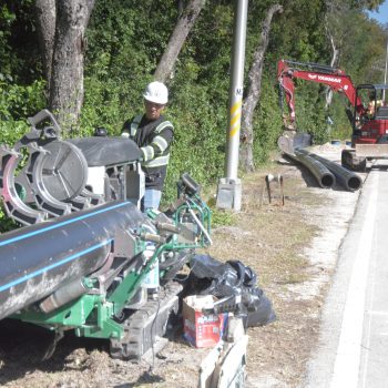 A construction worker in a safety vest and helmet operates machinery to join large black pipes beside a road, with an excavator and more pipes in the background near a wooded area.