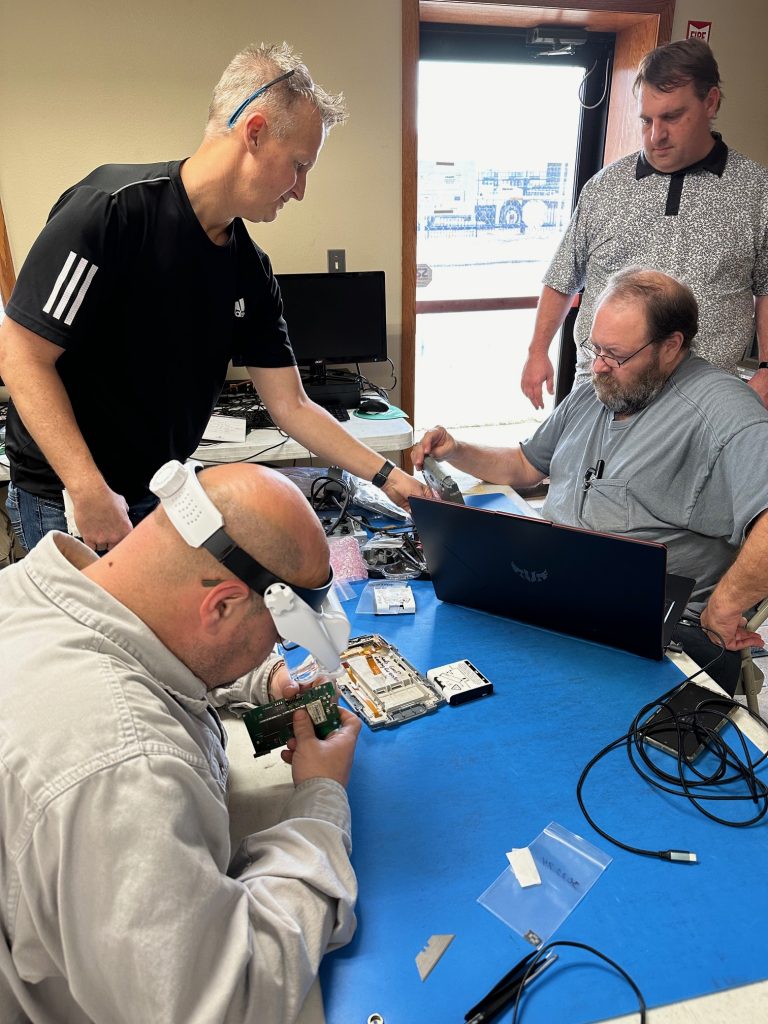 Four men work together at a table with electronics and computer parts. One man uses a magnifying headset to examine a circuit board, while others assist and observe. Laptops, cables, and tools are scattered on the table.