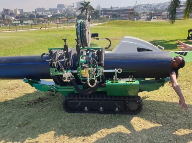 A woman playfully poses with her upper body inside a large, horizontal pipe being held by a green, tracked industrial machine outdoors on a grassy field. A cityscape is visible in the background.