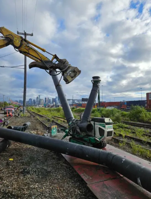 A construction digger lifts a large black pipe over a railway track, with a city skyline and scattered clouds visible in the background. Other pipes and construction materials are on the ground nearby.