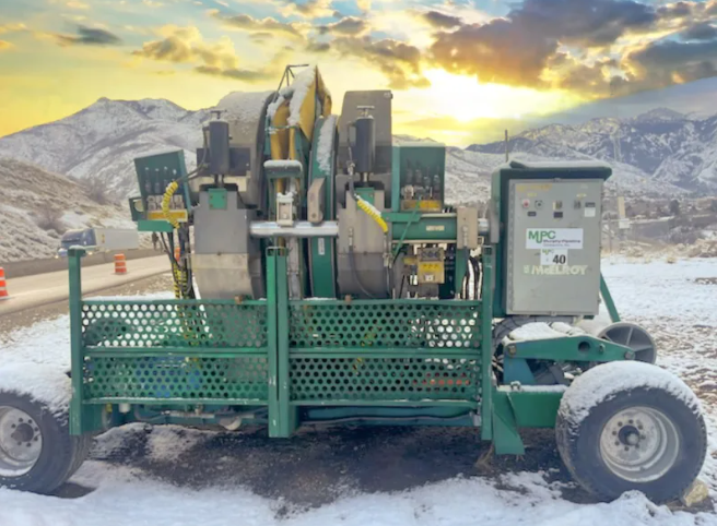 A green industrial machine with large wheels sits outdoors in a snowy landscape, mountains in the background, and a colorful sunrise sky overhead. Snow lightly covers the equipment and ground.