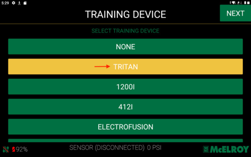 A digital interface titled Training Device shows a list of selectable devices. TRITAN is highlighted with a red arrow. Other options include NONE, 1200I, 412I, and ELECTROFUSION. A NEXT button is in the top right corner.