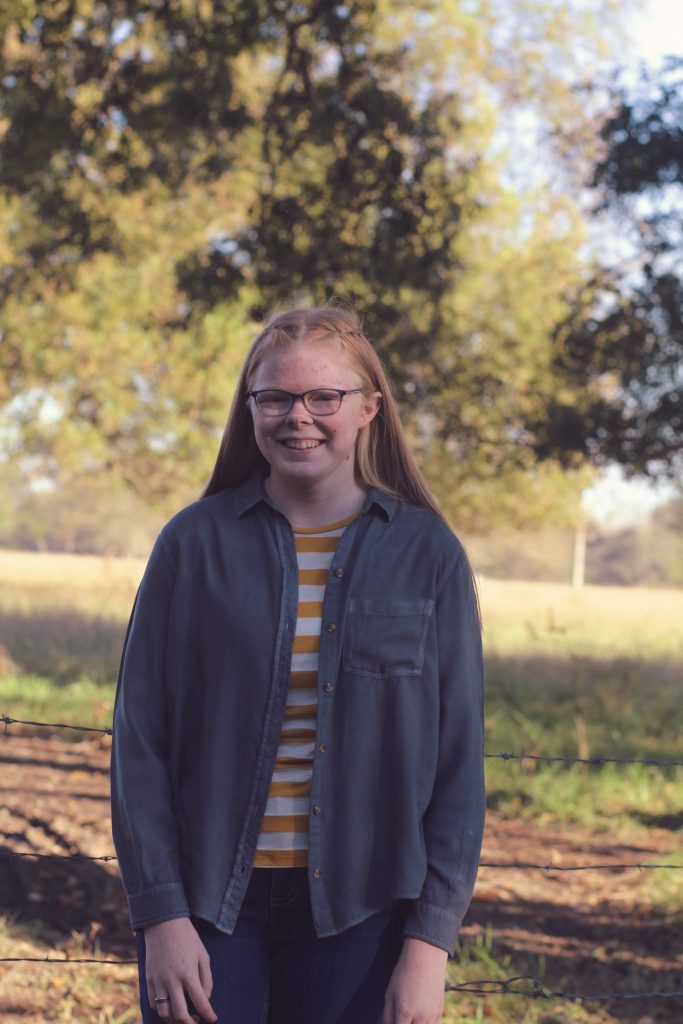 A young person with long straight hair and glasses smiles while standing outdoors in front of a fence, wearing a striped yellow and white shirt under an unbuttoned gray shirt, with trees and fields in the background.