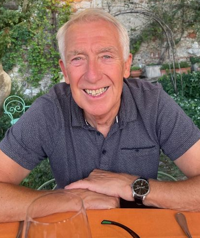 An older man with short gray hair smiles at a table outdoors, wearing a navy patterned polo shirt and a watch. Green plants and a stone wall are in the background. A glass and sunglasses are on the orange tablecloth.