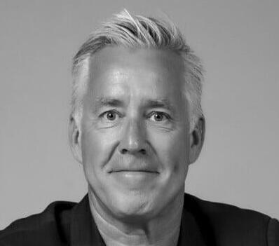 Black and white portrait of a middle-aged man with light skin and short, gray hair, smiling gently and looking directly at the camera. He is wearing a dark jacket against a plain background.