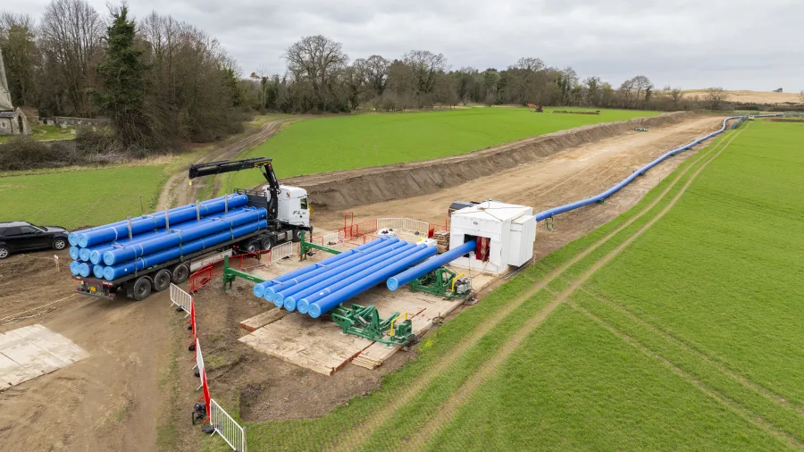 A construction site in a green field with a truck carrying large blue pipes, machinery positioning the pipes, and a long trench with blue pipes laid out, under a cloudy sky.