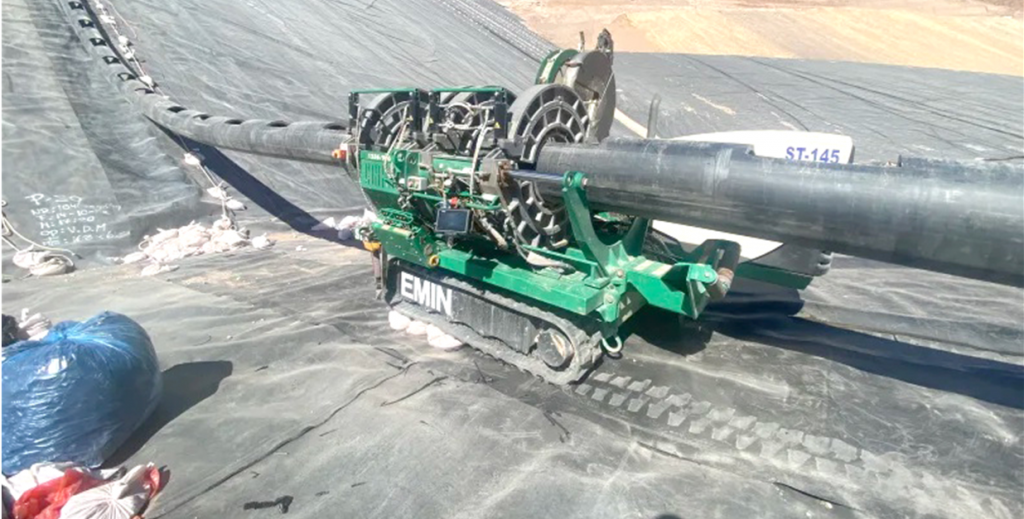 A green pipe fusion machine is welding large black pipes on a sloped surface covered with black geomembrane at a construction or industrial site. Equipment and materials are visible nearby.