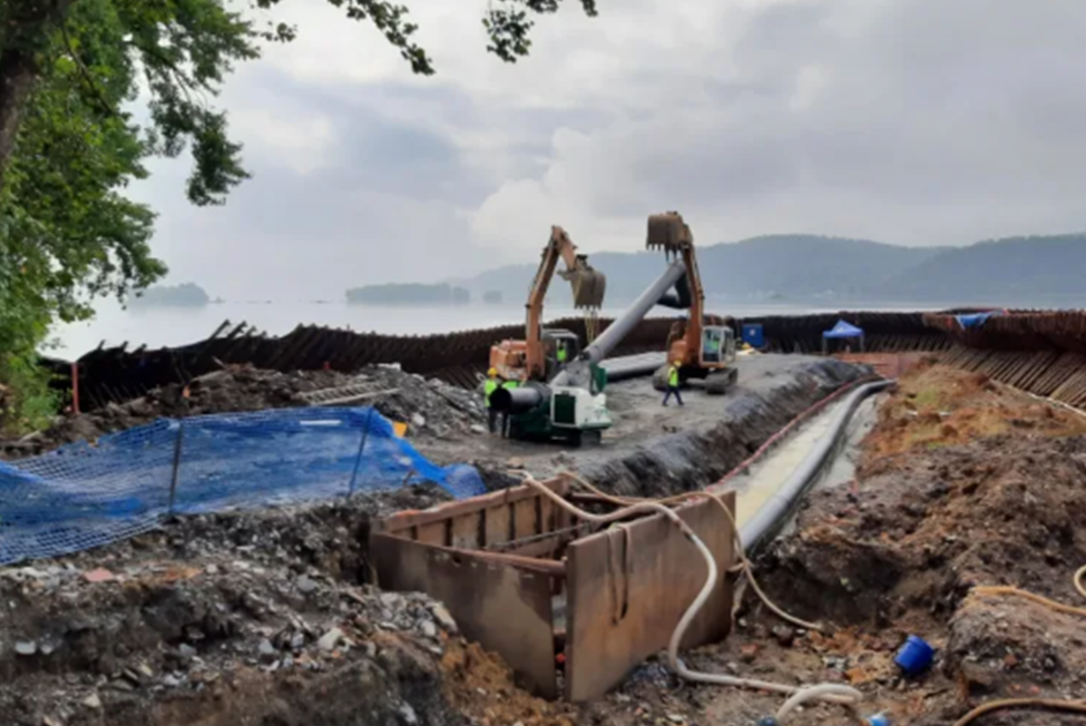 Two excavators lift and position a large pipe at a construction site near a body of water, with workers in safety gear nearby and hills visible in the background under a cloudy sky.
