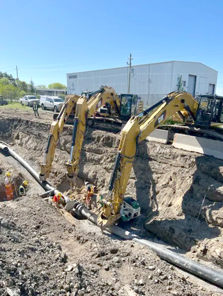 Three large excavators lift and position a pipeline in a deep trench while several construction workers in safety gear assist at the site. A white industrial building is visible in the background under a clear sky.