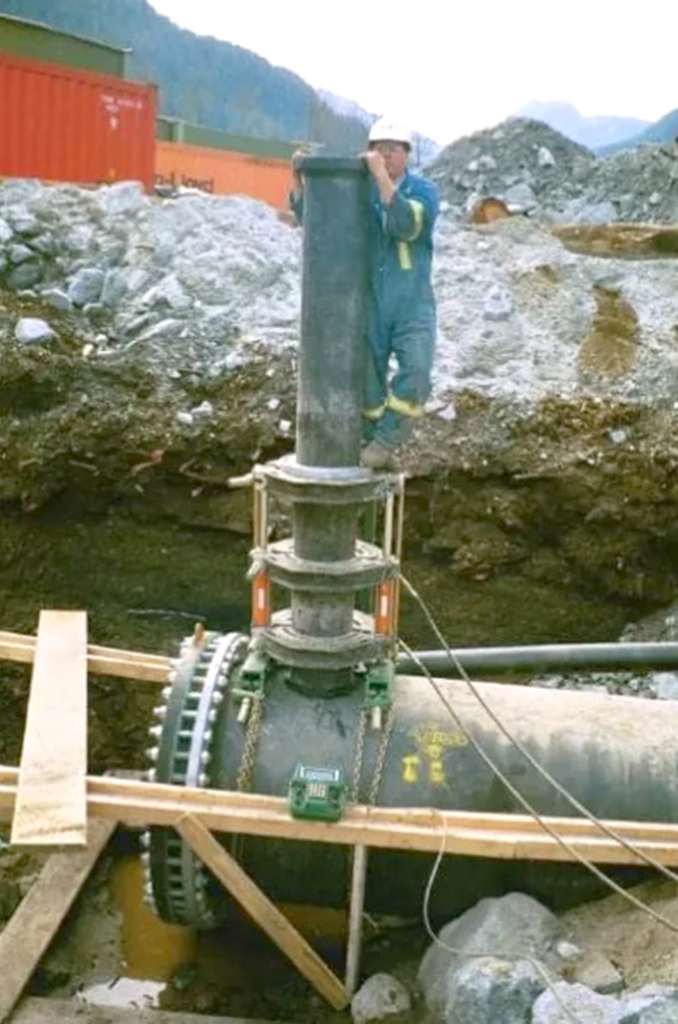 A worker in safety gear stands next to a large valve being installed on an underground pipeline at a construction site, surrounded by dirt, rocks, wooden supports, and equipment.