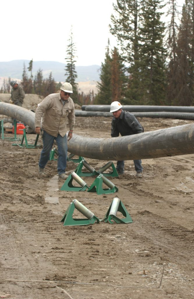 Two workers in hard hats guiding a large pipeline into place on green supports at a muddy, outdoor construction site surrounded by trees.