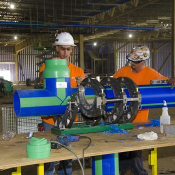 Two construction workers in orange shirts and hard hats work together on assembling large blue and green pipes inside a warehouse, using specialized equipment on a workbench cluttered with tools and materials.